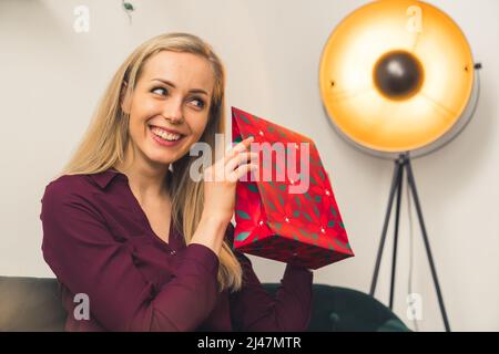 Es ist an der Zeit, Geschenke zu eröffnen - ein attraktives blondes europäisches Mädchen mit einem medialen Nahaufnahme-Studio, das ein Geschenk und ein Lächeln im Gesicht hält. Hochwertige Fotos Stockfoto