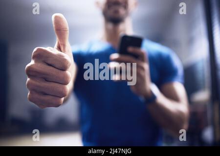 Dieser persönliche Trainer genehmigt diese App. Aufnahme eines nicht erkennbaren Mannes, der mit einem Smartphone zeigt und in einem Fitnessstudio Daumen nach oben zeigt. Stockfoto