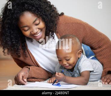 Welches Meisterwerk erschaffen Sie jetzt? Aufnahme einer attraktiven jungen Mutter, die mit ihrem Sohn auf dem Boden des Wohnzimmers liegt und ihm beim Zeichnen zuschaut. Stockfoto