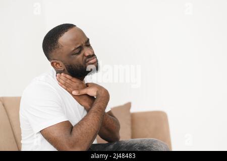 Schreckliche Halsschmerzen eines schwarzen Mannes. Hustet einen hübschen jungen afroamerikanischen Mann, der seinen Hals berührt, während er zu Hause auf dem Sofa sitzt. Wird krank. Stockfoto