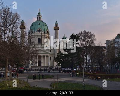 Blick über den öffentlichen Park Resselpark im historischen Zentrum von Wien, Österreich mit der berühmten Karlskirche und demonstrierenden Menschen am Abend. Stockfoto