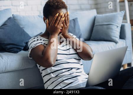 afroamerikanische Frau, die mit einem Laptop auf dem Boden saß, stresste das Gesicht mit Handflächen. Frau, die an einem Laptop arbeitet. Trauriges Gesicht. Stockfoto