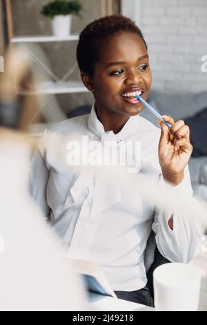 Lächelnde, verträumte afroamerikanische Frau, Projektarbeit am Arbeitsplatz. Moderne Kommunikation. Unternehmensporträt. Modernes Design. Stockfoto