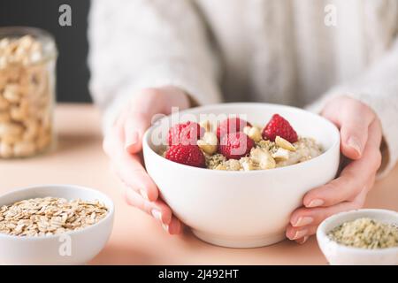 Haferflockenschale mit Himbeere, Cashewnüssen und Hanfsamen in den Händen der Frau. Sauberes Essen, Diät-Konzept Stockfoto