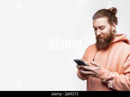 Skandinavischer, gutaussehender, glücklicher Mann, der mit kabellosen Kopfhörern in orangefarbenem Sweatshirt auf grauem Hintergrund telefoniert und lächelt. Kerl mit Ingwer h Stockfoto