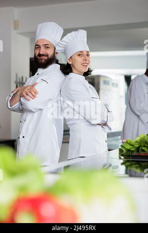 Erfahrene Köche tragen Kochuniformen, während sie sich im Küchenbereich des Restaurants von hinten auflehnen. Multiethnische Gastronomieexperten stehen in gekreuzten Armen und lächeln vor der Kamera. Stockfoto