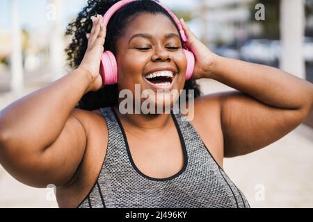 Kurvige Frau tanzt und hört Musik mit Kopfhörern beim Joggen im Freien - Fokus auf Gesicht Stockfoto