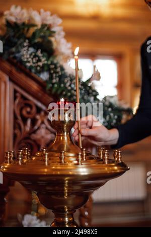 Zugeschnittenes Foto einer Frau, die in der orthodoxen Kirche brennende Kerze auf einen goldenen Ständer mit Kerzenhaltern in der Nähe von weißen Blumen setzt. Stockfoto