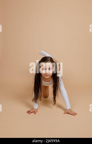 Portrait eines jungen niedlichen konzentrierten Mädchens mit langen dunklen Haaren in weißem Trainingsanzug und Socken, die auf allen Vieren stehen und das Bein hochheben und sich darauf posieren Stockfoto