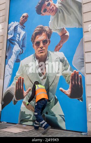 Ein Straßenputzer nimmt am 11.. April 2022 in London, England, Karton auf, den die Homeles hinterlassen haben, unter einer Plakatwand für einen Modehändler im Zentrum von London. Stockfoto