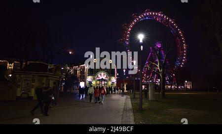 Nachtansicht von Menschen, die zum Eingang des Vergnügungsparks Wurstelprater in Wien, Österreich, mit dem berühmten erillten Riesenrad laufen. Stockfoto