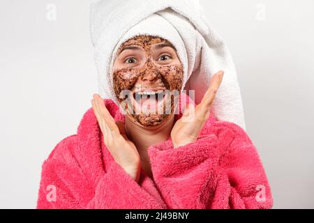 Eine Frau mit einer Maske auf ihrem Gesicht in einer rosa Robe und einem Handtuch auf ihrem Kopf schreit und bewundert den weißen Hintergrund Stockfoto