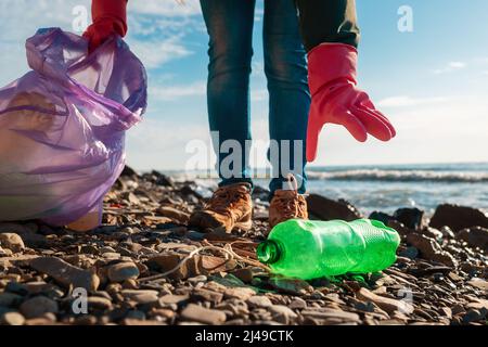 Ein Freiwilliger in Gummihandschuhen greift nach einer schmutzigen Plastikflasche, die am Meeresufer liegt. Nahaufnahme der Hand. Das Konzept der Reinigung des wilden Strandes. Stockfoto