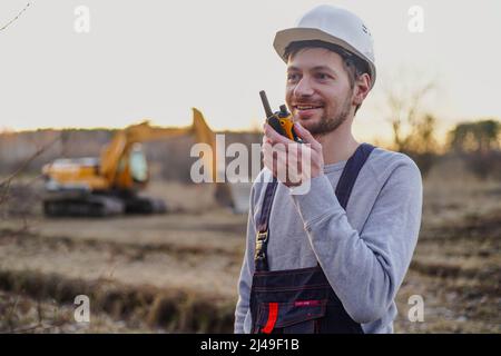 Lächelnder Baumeister im Helm, der durch den Funksender auf der Baustelle spricht. Bagger im Hintergrund. Stockfoto