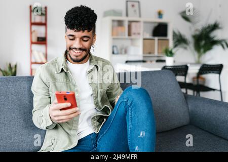 Multirassischer Mann, der auf einer Couch im Wohnzimmer sitzt und ein Mobiltelefon benutzt Stockfoto