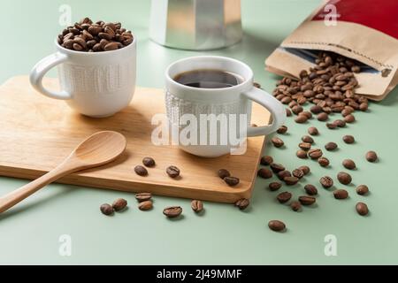 Tasse frisch gebrühten Kaffee, Löffel und verstreute Kaffeebohnen auf pastellgrünem Hintergrund. Aus einem braunen Papierbeutel verschüttete Bio-Kaffeebohnen. Stockfoto