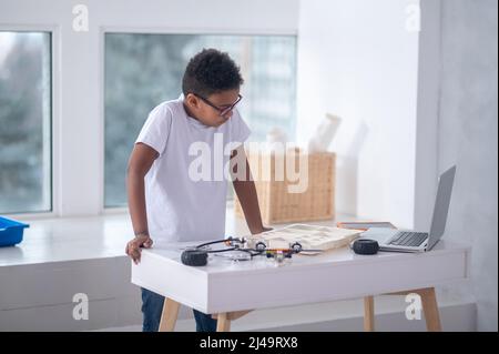 Ein Junge in einem weißen T-Shirt, der mit Kabeln und Laptop am Tisch steht Stockfoto