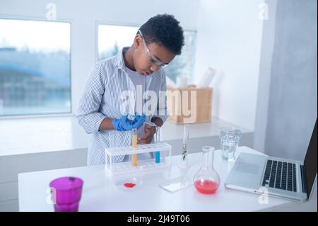 Ein dunkelhaariger Junge in einem Labormantel, der während eines chemischen Experiments beschäftigt aussieht Stockfoto