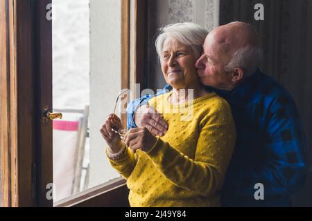 Älteres kaukasisches Paar am Fenster. Glückliche Beziehung. Der alte kahle Mann umarmte seine Frau und gab ihr einen Kuss auf ihr Küken. Hochwertige Fotos Stockfoto