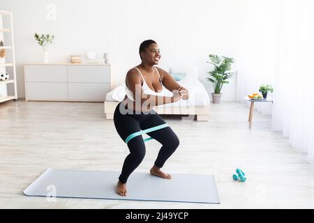 Mollige tausendjährige schwarze Frau, die während des häuslichen Trainings mit Gummiband hockt, freier Platz Stockfoto