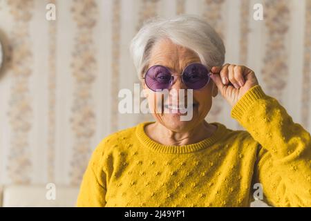 Funky lustige grauhaarige kaukasische Großmutter im Ruhestand, die die Kamera ansah und lächelte, während sie eine moderne violette Brille trug. Mittlere Innenaufnahme mit Vintage-Tapete. Hochwertige Fotos Stockfoto