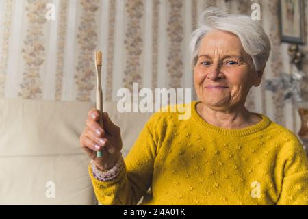 Senioren wählen Ökologie. Freundliche, glückliche kaukasische, grauhaarige Großmutter, die eine ökologische Bambuszahnbürste in der Hand hält und die Kamera anschaut. Hochwertige Fotos Stockfoto