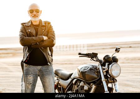 Auffälliger älterer Mann in Lederjacke, der am Sommertag mit dem Motorrad im Freien posiert Stockfoto