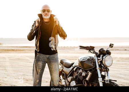 Auffälliger älterer Mann in Lederjacke, der am Sommertag mit dem Motorrad im Freien posiert Stockfoto