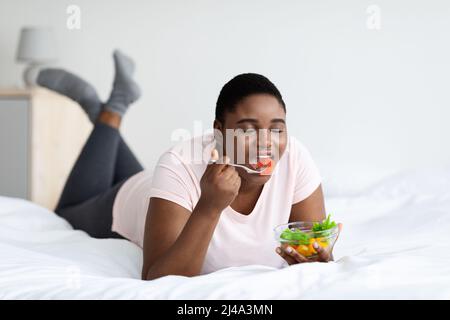 Übergewichtige schwarze Frau, die leckeren Gemüsesalat isst und zu Hause auf dem Bett liegt. Gesunde Ernährung zur Gewichtsabnahme Stockfoto