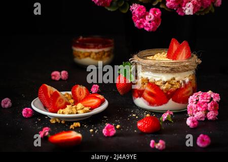 Erdbeerdessert auf schwarzem Hintergrund. Tiramisu und rosa Blüten. Erdbeeren mit Rahm und Keksen in einem Glas. Stockfoto