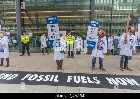 London, Großbritannien. April 2022. Eine Gruppe von Wissenschaftlern und medizinischen Fachleuten in weißen Labormänteln protestiert gegen die Entwicklung fossiler Brennstoffe außerhalb eines modernen Gebäudes mit Glasfassade. Sie halten Plakate mit der Aufschrift „WISSENSCHAFT SAGT NEUES ÖL & GAS = TOD“ und stehen hinter einem großen Banner mit der Aufschrift „ENDE FOSSILER BRENNSTOFFE JETZT“. Polizeibeamte überwachen die friedliche Demonstration und betonen die Dringlichkeit und den wissenschaftlichen Konsens zum Klimaschutz. Wissenschaftler der Extinction Rebellion kleben ihre Hände an die Fenster vor dem Department for Business, Energy & Industrial Strategy in der Victoria Street. Penelope Barritt/Alamy Stockfoto
