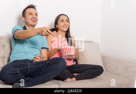 Latino-Mann und -Frau, die mit der Fernbedienung Fernsehen und Popcorn essen, die zu Hause auf einer Couch sitzen Stockfoto