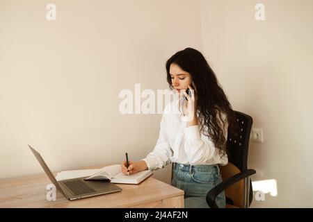 Eine vielbeschäftigte Frau spricht per Telefon, schreibt Notizen auf ein Notebook und arbeitet am Laptop. Müde Mädchen Chef arbeitet online mit Kollegen zu Hause Stockfoto