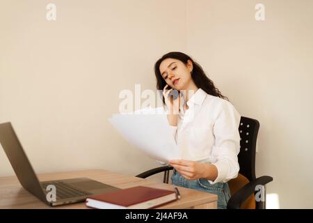 Fröhliche Managerin, die telefonisch spricht, Schreibarbeiten macht und am Laptop arbeitet. Mädchen Chef arbeiten im Büro Stockfoto