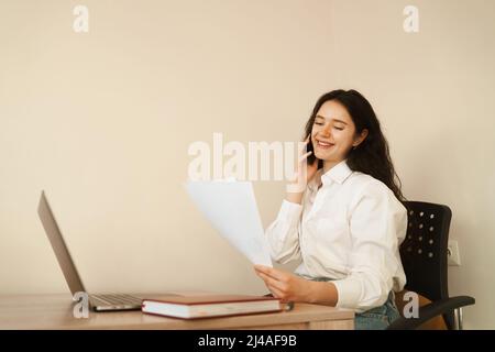 Fröhliche Managerin, die telefonisch spricht, Schreibarbeiten macht und am Laptop arbeitet. Mädchen Chef arbeiten im Büro Stockfoto