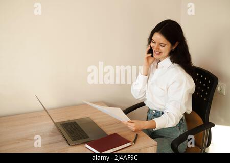 Fröhliche Managerin, die telefonisch spricht, Schreibarbeiten macht und am Laptop arbeitet. Mädchen Chef arbeiten im Büro Stockfoto