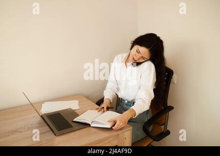 Eine vielbeschäftigte Frau spricht per Telefon, schreibt Notizen auf ein Notebook und arbeitet am Laptop. Müde Mädchen Chef arbeitet online mit Kollegen zu Hause Stockfoto