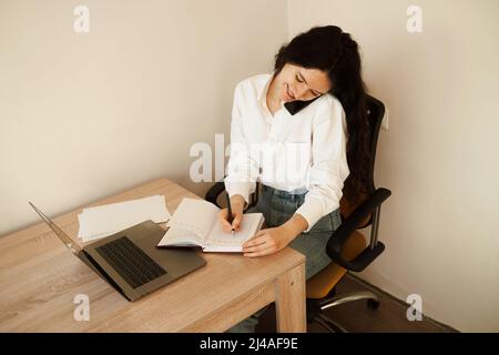 Eine vielbeschäftigte Frau spricht per Telefon, schreibt Notizen auf ein Notebook und arbeitet am Laptop. Müde Mädchen Chef arbeitet online mit Kollegen zu Hause Stockfoto