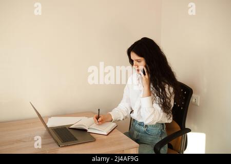 Eine vielbeschäftigte Frau spricht per Telefon, schreibt Notizen auf ein Notebook und arbeitet am Laptop. Müde Mädchen Chef arbeitet online mit Kollegen zu Hause Stockfoto