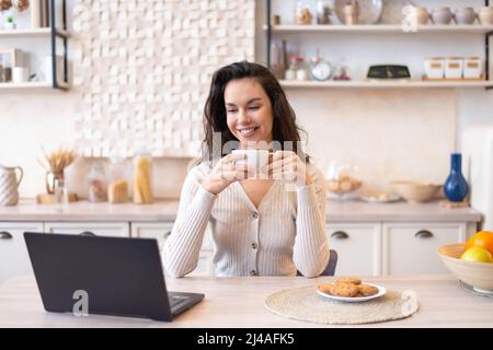 Fröhliche Frau, die vor dem Laptop am Küchentisch sitzt, am Computer Filme guckt und Kaffee trinkt Stockfoto