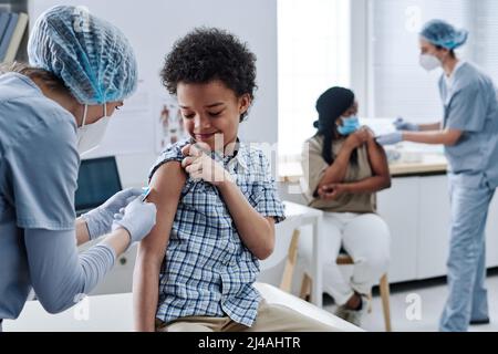 Afrikanischer tapferer Junge, der während des Eingriffs im Krankenhaus mit anderen Personen im Hintergrund von der Krankenschwester eine Impfung in der Schulter erhält Stockfoto