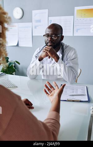 Afrikanischer männlicher Arzt sitzt am Tisch in seinem Büro und hört dem Patienten während des Besuchs aufmerksam zu Stockfoto