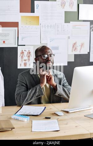 Afrikanischer Arzt, der am Tisch vor dem Computermonitor sitzt und online mit dem Patienten im Büro spricht Stockfoto
