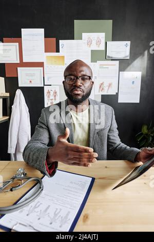 Afrikanischer Facharzt für Brillen, der medizinische Karten und Dokumente verwendet, während er während einer Online-Konferenz am Computer im Büro spricht Stockfoto