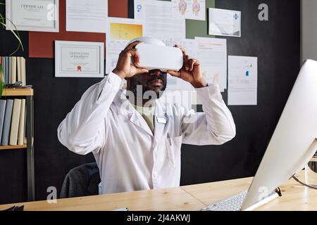 Afrikanischer männlicher Arzt im weißen Mantel sitzt an seinem Arbeitsplatz mit Virtual-Reality-Brille spielen Videospiel Stockfoto