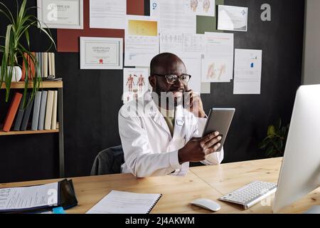Afrikanischer männlicher Arzt, der am Arbeitsplatz sitzt und Tablet-pc hält und lächelt, während er auf dem Mobiltelefon spricht Stockfoto