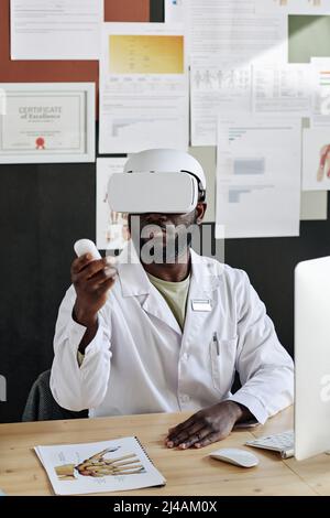 Afrikanischer männlicher Arzt in Drahtlosbrille, der virtuelle Realität spielt, während er am Tisch im Büro sitzt Stockfoto