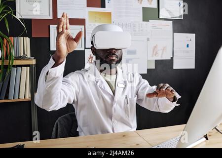 Afrikanischer Arzt in einer virtuellen Brille, der in der Luft gestikuliert, während er an seinem Arbeitsplatz im Büro sitzt Stockfoto