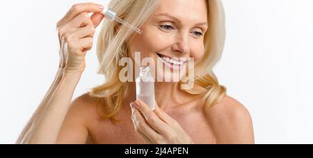 Eine hochentwickelte Frau mittleren Alters trägt regenerierendes Serum auf das Gesicht auf. Charmante blonde 40s Dame mit Anti-Aging-Komplex mit Hyaluronsäure isoliert auf weißem Hintergrund. Hautpflegekonzept Stockfoto