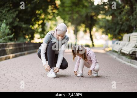 Familienwanderung und Fitsporttraining. Die angenehme junge arabische Mutter schnürt Schuhe, während ihre Tochter ihre Turnschuhe anpasst und am Sommermorgen auf der Joggingstrecke im Park sitzt. Stockfoto